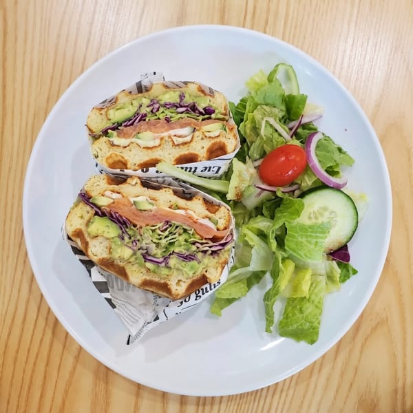 Two waffle sandwiches with avocado, salmon, and cabbage, served with a side salad on a white plate.