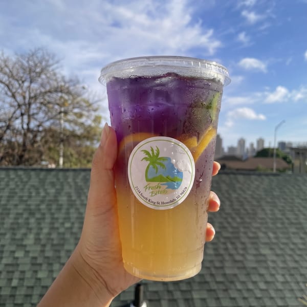 Hand holding a Fresh Bites drink with purple and yellow layers, against a blue sky.