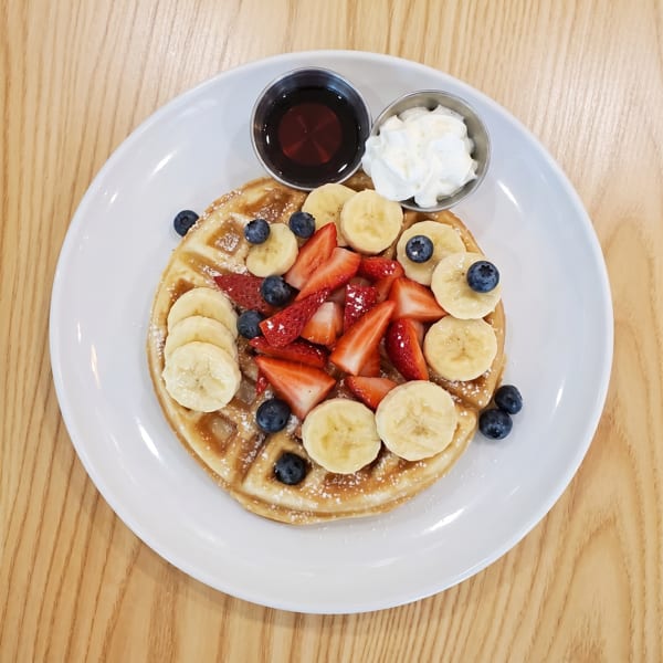 A golden waffle topped with sliced bananas, strawberries, blueberries, powdered sugar, and sides of syrup and whipped cream.