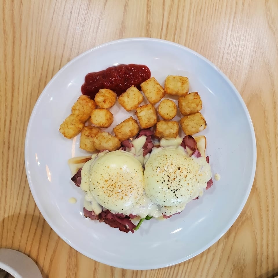 Eggs Benedict with pastrami, tater tots, and ketchup on a white plate.